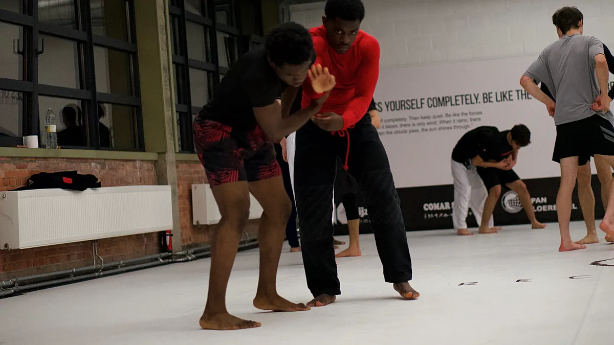 Twee BJJ-beoefenaars oefenen een grappling techniek in de guard positie tijdens een groepsles in een trainingshal.
