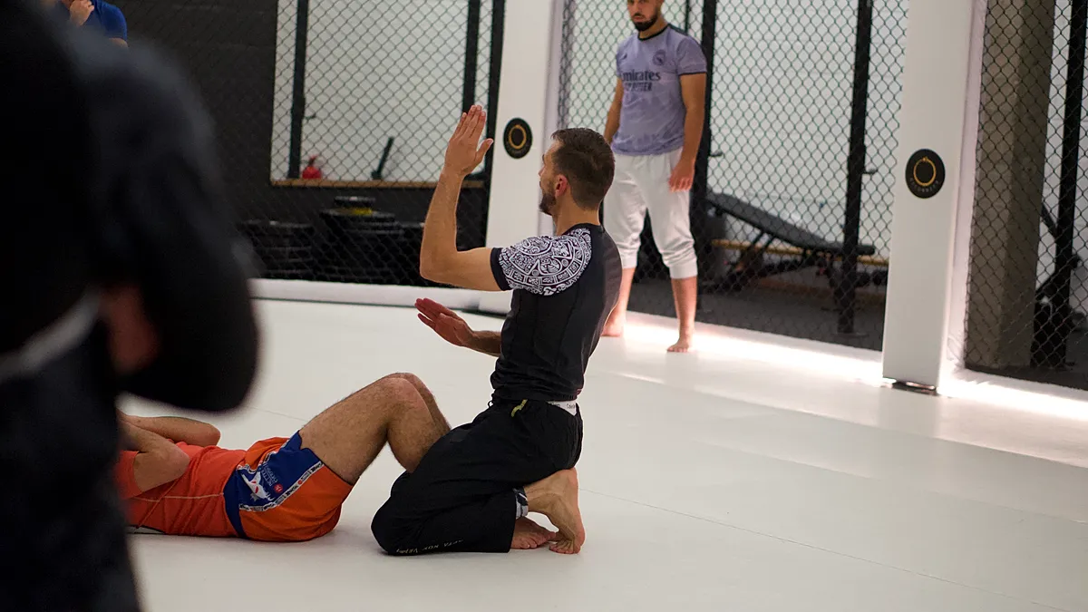 Een BJJ-beoefenaar werkt aan guard-positie terwijl trainers en medestudenten observeren in de trainingsruimte.