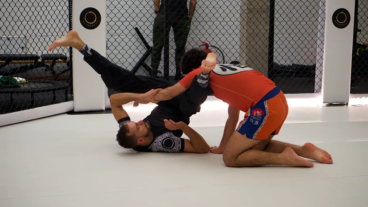 Twee BJJ-beoefenaars trainen op de mat waarbij één fighter in guard positie ligt en de ander een sweep-beweging uitvoert.