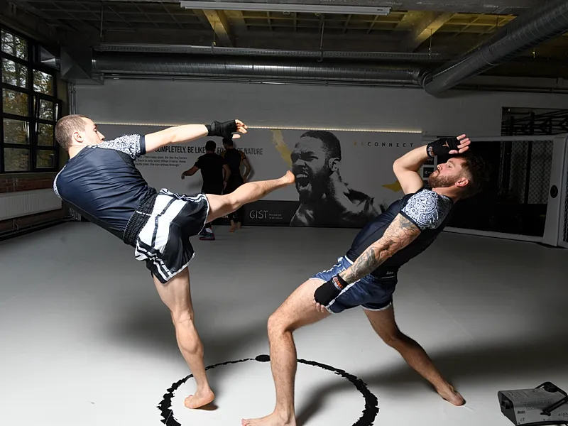 Twee MMA-atleten voeren kickboksings- en slagwerk uit met pads in een trainingsgymnasium.