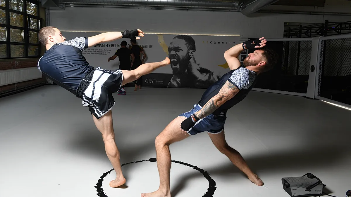 Twee MMA-atleten voeren kickboksings- en slagwerk uit met pads in een trainingsgymnasium.