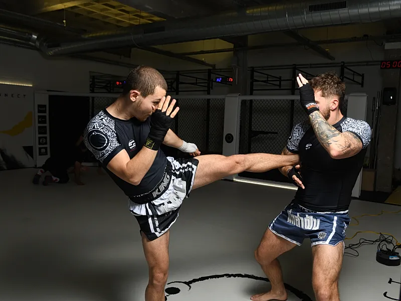 Twee MMA-fighters voeren kickboksing-oefeningen uit waarbij één fighter een hoge trap uitvoert terwijl de ander in verdedigende houding staat.